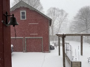 Red barn with snow on the ground and falling in the background.