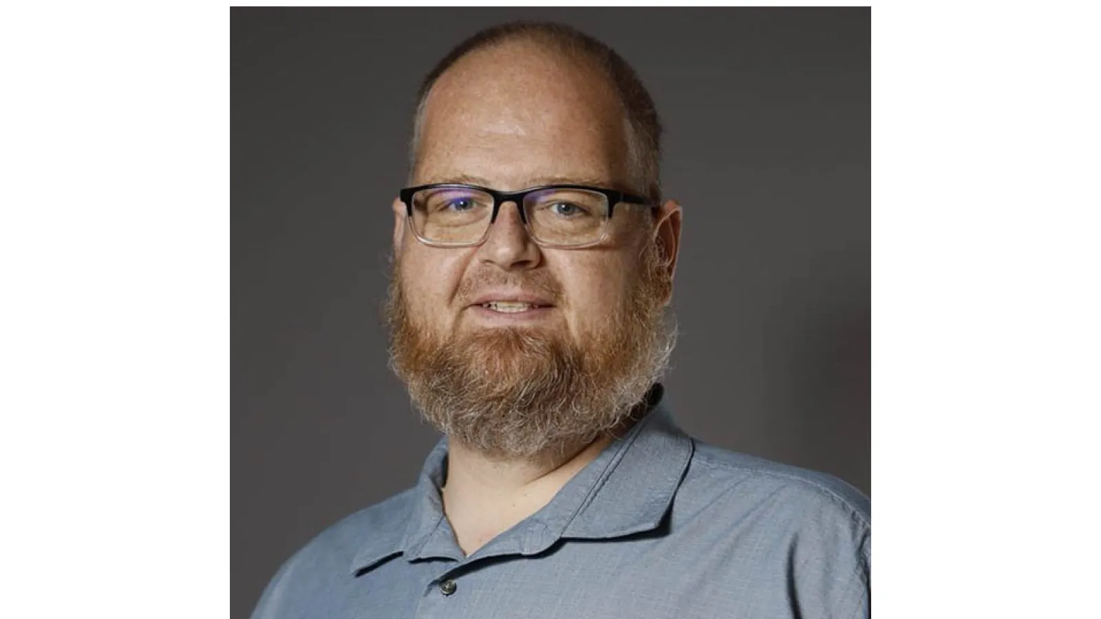 portrait of Aaron Wilson with light background wearing dark blue collar shirt and glassed with beard and short hair
