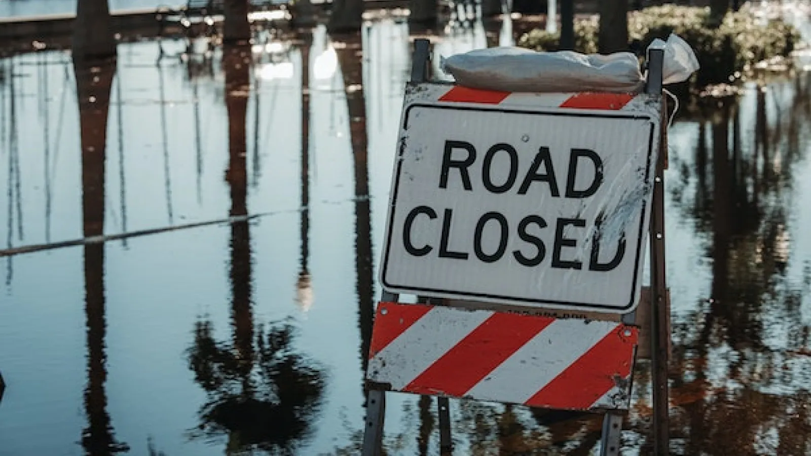 A Road Closed sign with a sandbag on it blocking a flooded road.