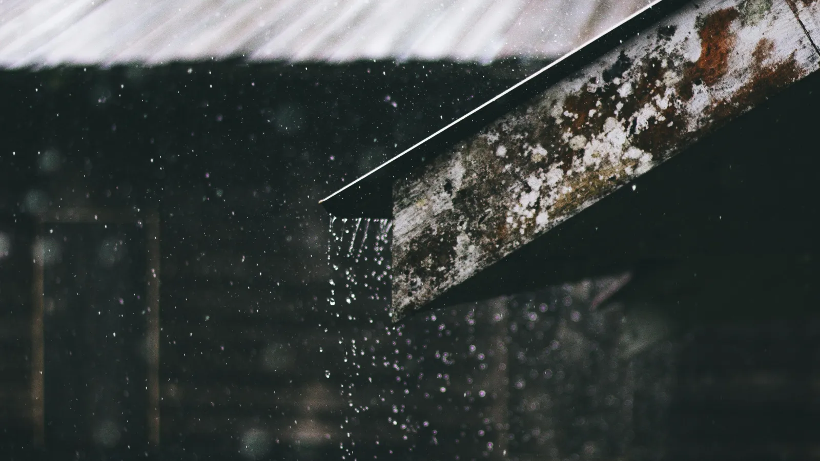 the side view of a slanted roof in the foreground with another metal roof in the background with rain falling and pouring down the roof in the foreground. 