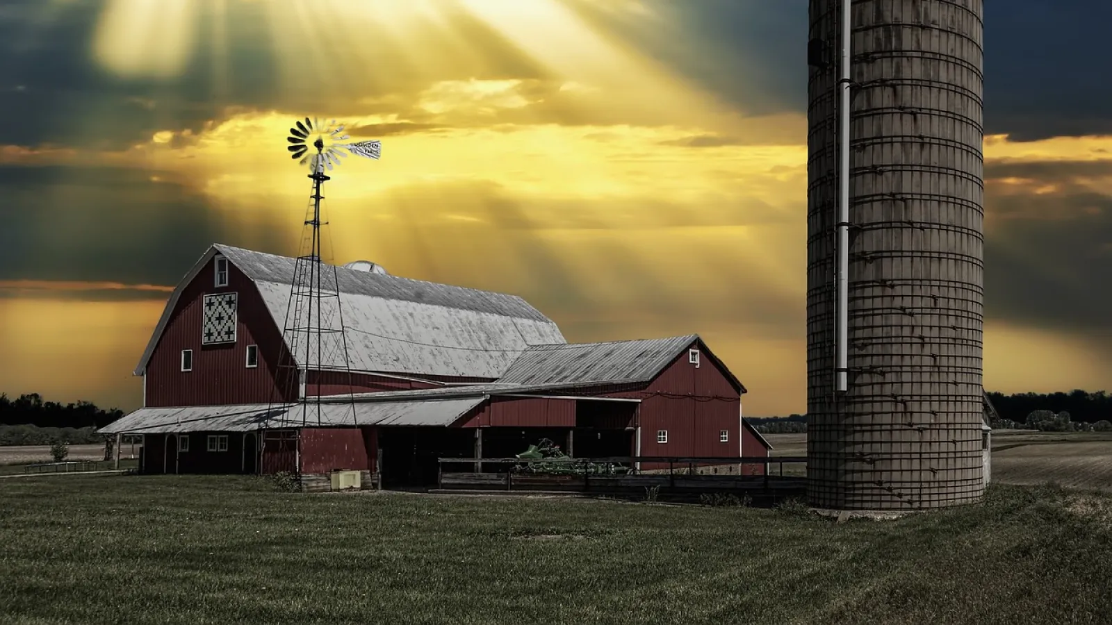 A farmhouse with a silo under a cloudy, dark sky with rays of sunshine coming through.
