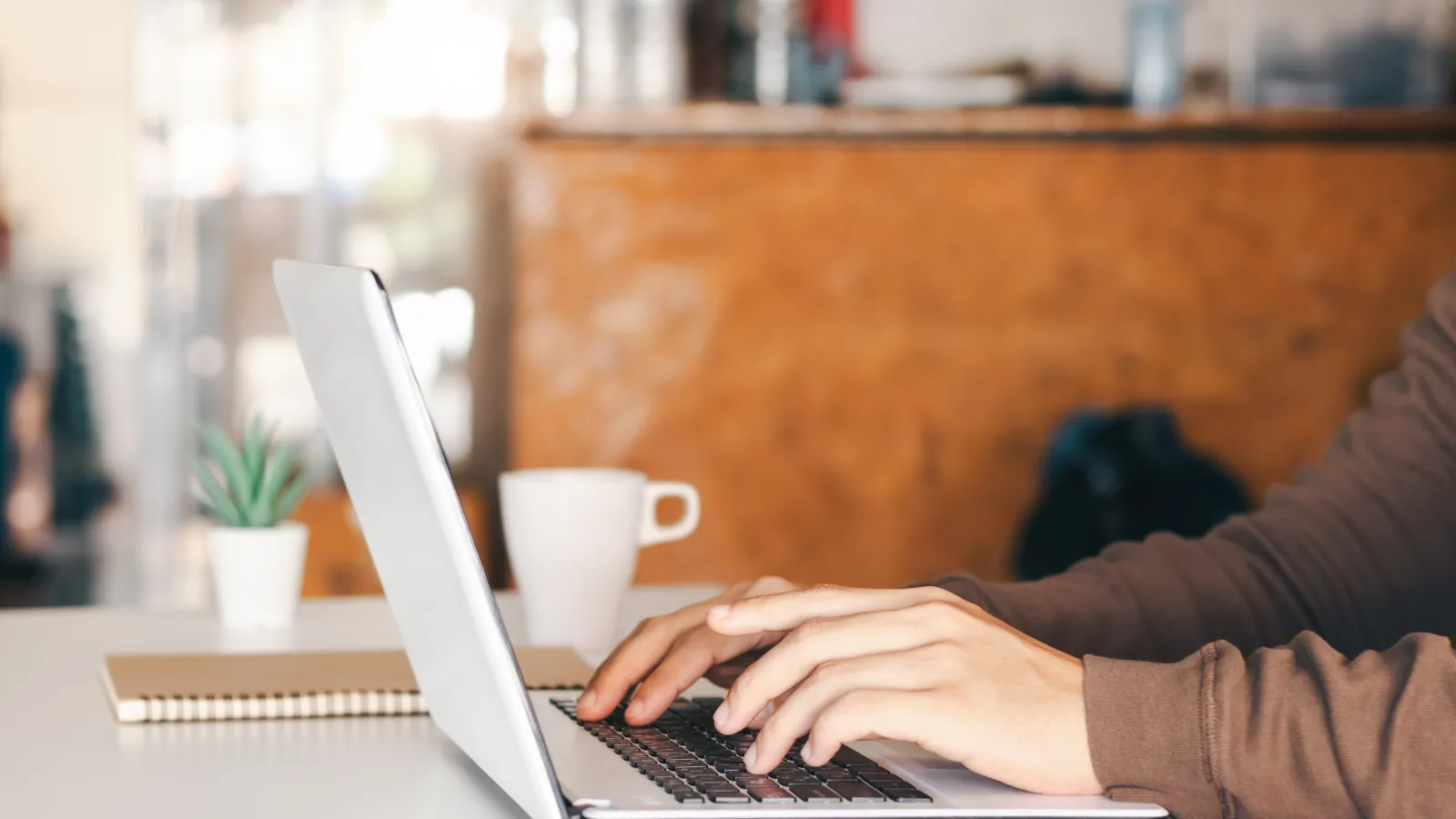 A person typing on a laptop on a wooden table, with a notebook and a coffee cup beside the laptop.