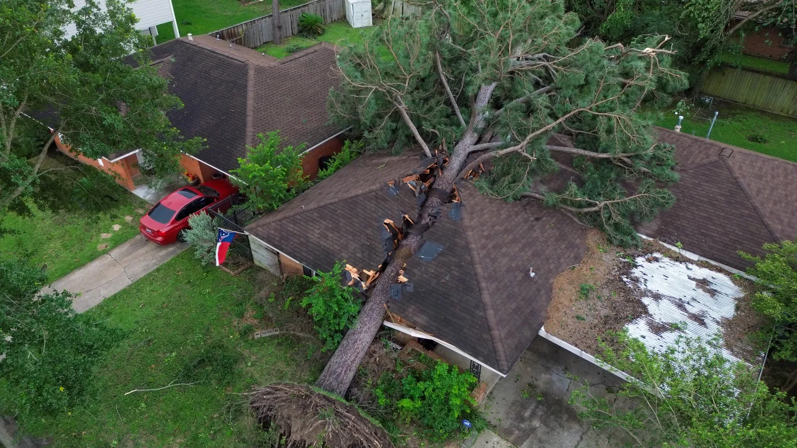 Aerial view of a large tree fallen on a house causing significant damage, with a red car parked nearby.