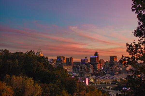 View from a hill among trees of downtown Cincinnati buildings during sunrise, blue-pink skies with some light feather white-pink clouds  