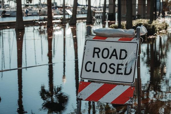 A Road Closed sign with a sandbag on it blocking a flooded road.