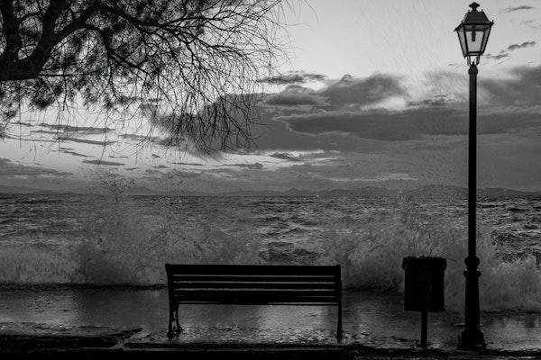 Black and white image of Park bench next to light post facing water getting flooded from water surge