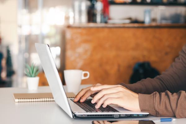 A person typing on a laptop on a wooden table, with a notebook and a coffee cup beside the laptop.